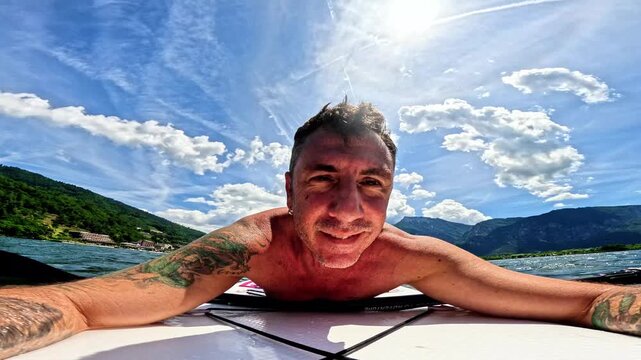 A man lying on a stand-up paddle board in Lake Levico, behind him, in addition to the blue sky with white clouds, the mountains overlooking the lake