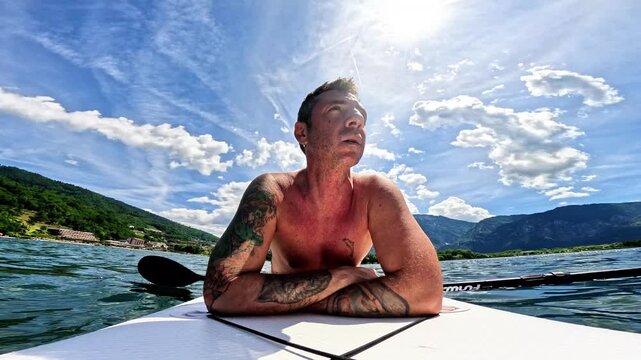 A man lying on a stand-up paddle board in Lake Levico, behind him, in addition to the blue sky with white clouds, the mountains overlooking the lake
