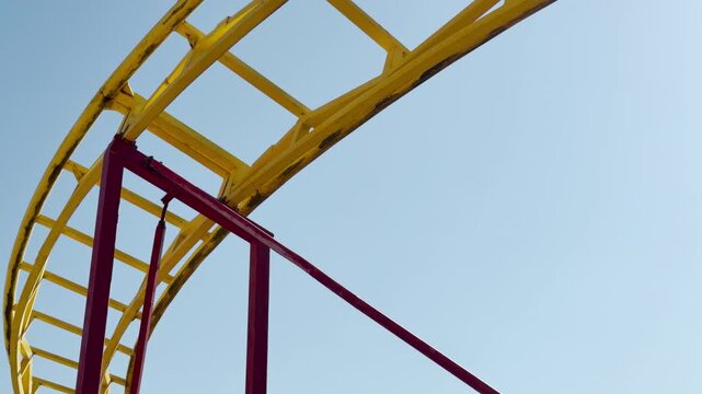 A vibrant yellow and red steel roller coaster track bends against a clear blue sky. The sight brings feelings of thrilling anticipation, nostalgic joy, and lighthearted summer fun.