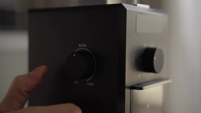 Close-up of hand turning adjustment dial on professional electric burr grinder, selecting desired consistency from coarse to fine for brewing aromatic coffee. Shooting in slow motion