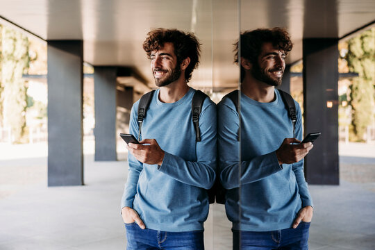 Smiling man with hand in pocket holding smart phone leaning on wall in corridor