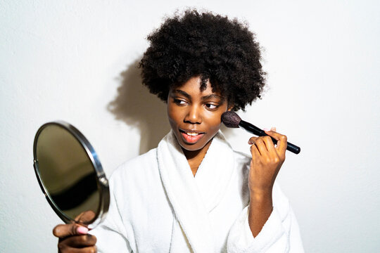 Curly hair woman in bathrobe doing make-up while standing against white background