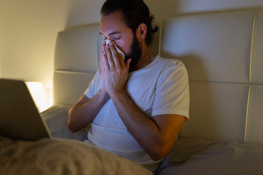 Man blowing nose and sitting with laptop on bed