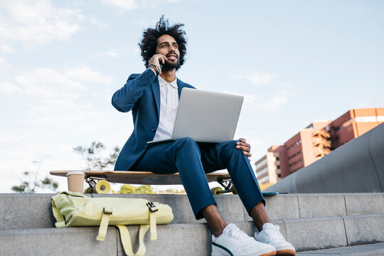 Spain, Barcelona, young businessman sitting outdoors in the city using cell phone and laptop