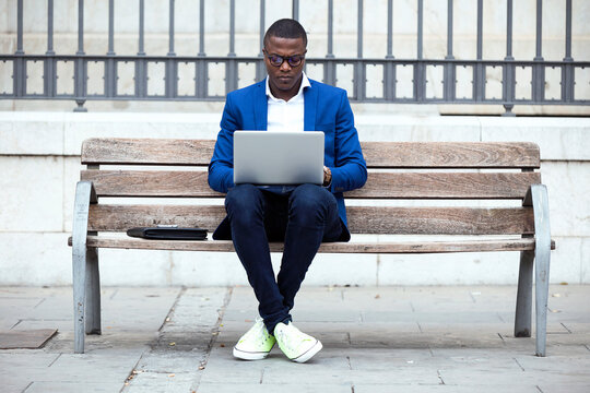 Young businessman wearing blue suit jacket sitting on bench and using laptop