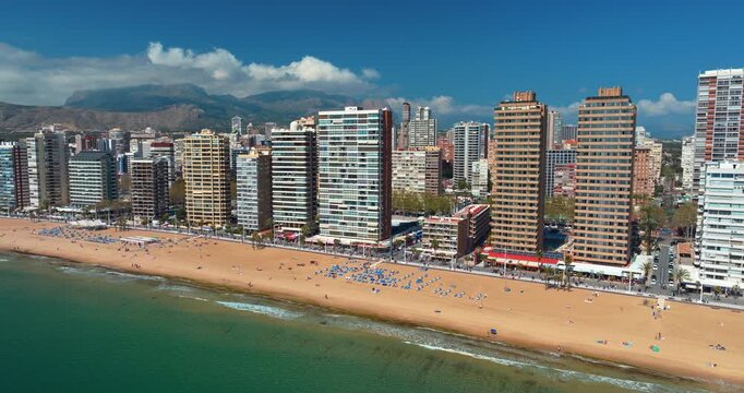Benidorm Levante beach aerial close view blue umbrellas crowd summer Spain Alicante seafront high-rise buildings Mediterranean coastline turquoise water mountain backdrop drone slow motion tourism