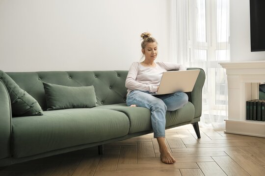 A woman sits comfortably on a green couch in a bright living room. She focuses on her laptop, dressed casually, surrounded by natural light streaming through large windows