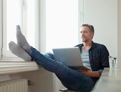 Germany, Cologne, Mature man sitting at window using laptop, feet up
