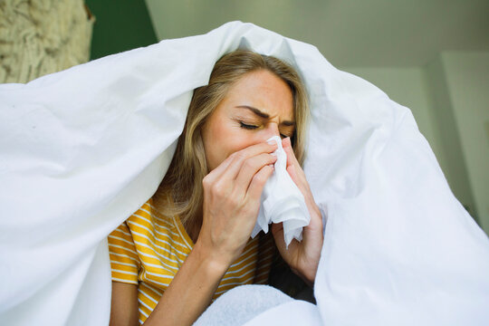 Woman wiping nose with tissue under blanket at home