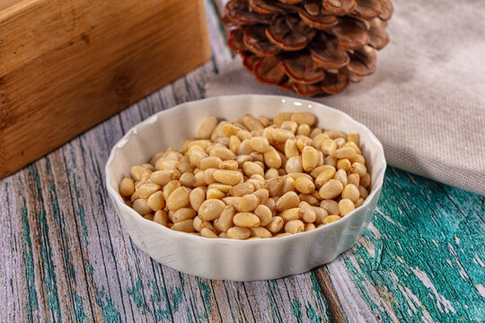 Pine nuts in a white bowl with a pine cone on a rustic wooden background