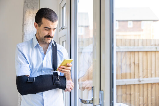 Man wearing arm sling using smart phone while standing by window at home