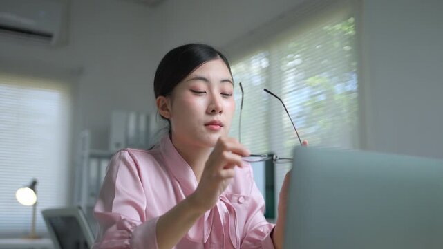 Exhausted Asian businesswoman taking off her glasses and massaging her tired eyes after working on the computer for a long time in the office