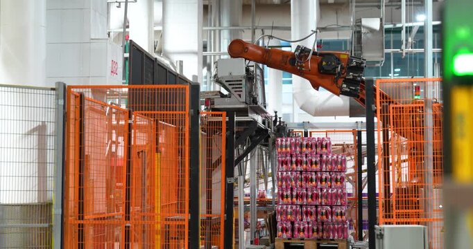 Steady shot of an automated orange industrial robotic arm palletizing packaged beverages behind a safety fence in a modern manufacturing plant