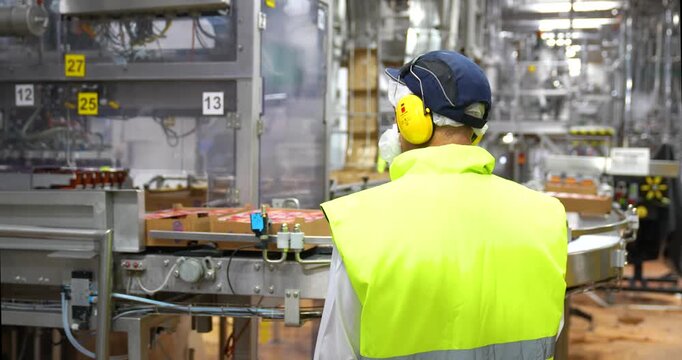 Steady over-the-shoulder shot of a factory worker in a high-visibility vest and ear protection monitoring an automated packaging line