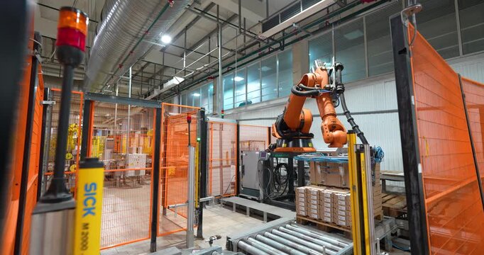 Steady wide shot of an automated orange industrial robotic arm palletizing cardboard boxes behind safety fences in a modern manufacturing plant