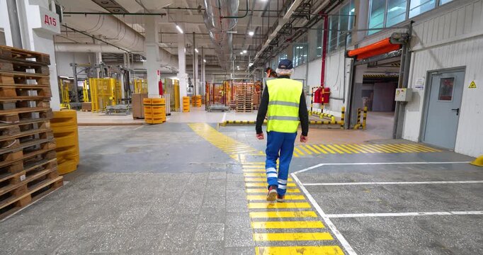 Cinematic tracking shot following a factory worker in a high-visibility vest walking along a marked yellow pedestrian path in a large industrial plant
