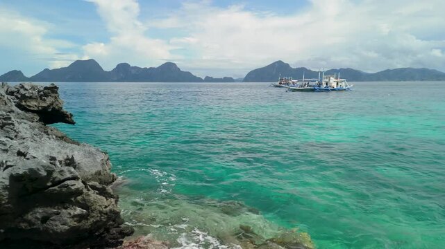 Outrigger tour boat with tourists arriving at Twin Rocks viewpoint by rugged limestone rock over clear turquoise sea with mountain backdrop in El Nido Palawan Philippines