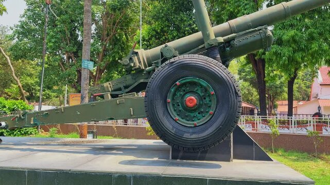 Cinematic side view panning shot of a 105 mm Indian Field Gun displayed on a monument platform with large wheels green military body and trees in bright daylight