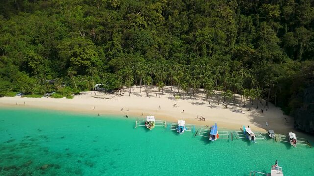 Aerial pullback rise revealing Seven Commandos Beach with tourist boats palm trees and turquoise water along forested coastline of El Nido Palawan Philippines