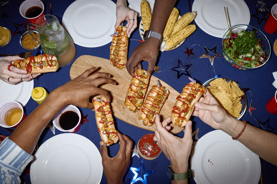 Top view shot of diverse young adults sharing hot dogs during Independence Day home gathering, celebrating holiday tradition and friendship. Ideal for holiday marketing and social campaigns