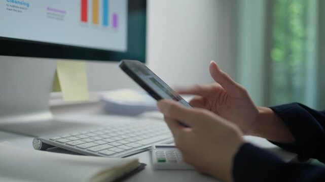 Close up of a businesswoman's hands scrolling and typing on a smartphone. An office setting with a computer monitor showing charts in the background