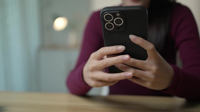Close up of a woman's hands holding and typing on a smartphone. She is sitting at a wooden table in a cozy home environment