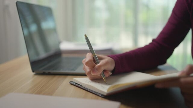 Close up of a female student's hand writing in a diary. A young businesswoman is taking notes on her agenda at a wooden desk with a laptop