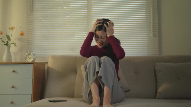 Frustrated asian woman sitting on the couch, feeling angry and stressed. Upset young lady screaming and experiencing a mental breakdown