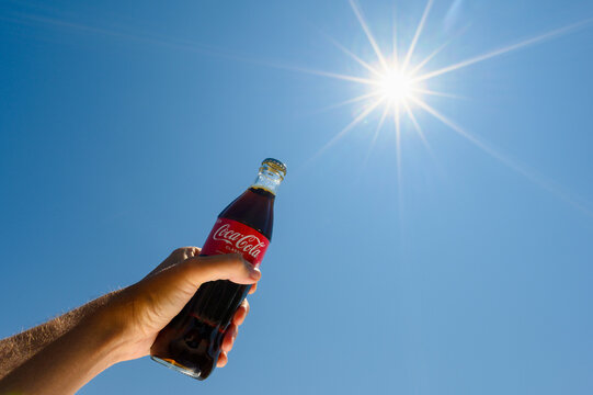 Anapa, Russia - July 30, 2023: A bottle of Coca-Cola in a man&rsquo;s hand against the background of the sky and bright sun