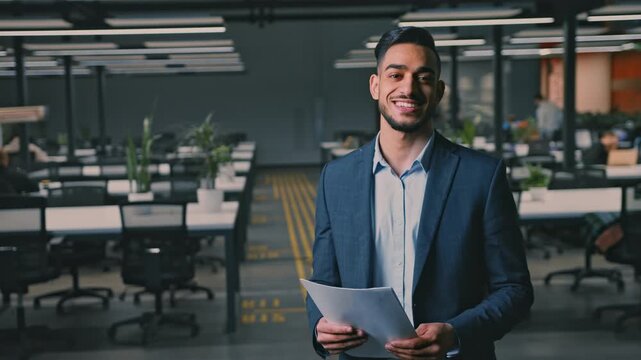 Focused Young Man Of Middle Eastern Descent Holding Documents While Standing In Modern Office, Engaged In Professional Activity. Stylish Workplace With Empty Desks And Chairs Around.