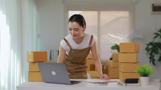 Young Asian woman running a small business from home, writing down order details while working on her laptop surrounded by cardboard boxes