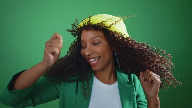 Expressive young woman with curly hair dancing and smiling happily in a studio. Stylish female model in a green blazer enjoying music and having fun while looking at the camera