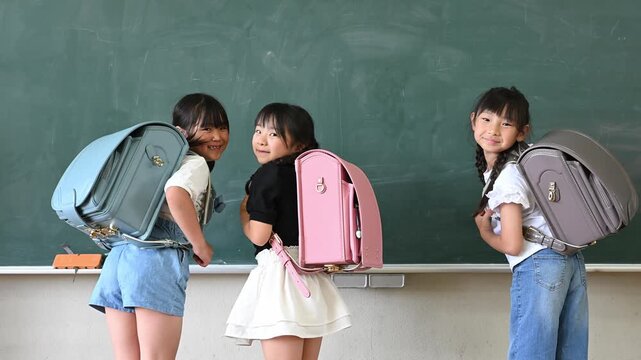 教室の黒板の前でカラフルなランドセルを背負い笑顔で振り返る新入生の女の子たち