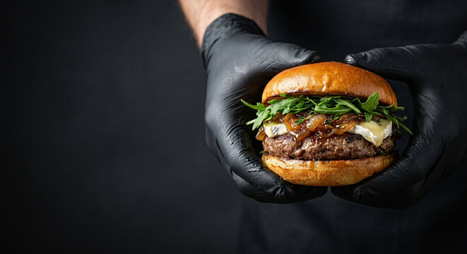 gourmet burger with thick beef patty, caramelized onions, melted Brie cheese, held in black gloves
