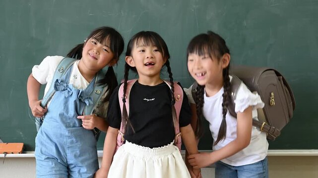 教室の黒板の前でカラフルなランドセルを背負い手をつなぐ笑顔の女子小学生たち