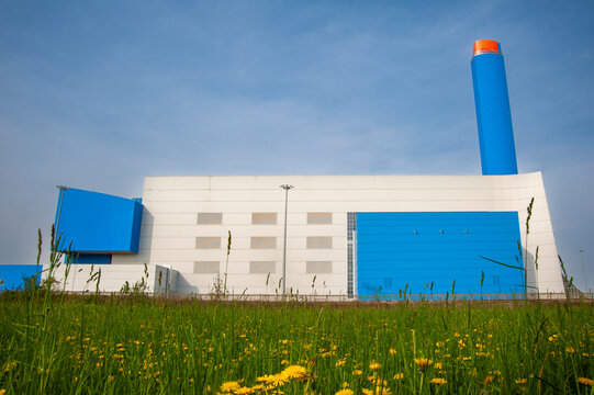 Struttura industriale moderna con pannelli blu, camino cilindrico e prato fiorito in primo piano. Ideale per concetti di architettura industriale, energia, sostenibilit&agrave; e infrastrutture.