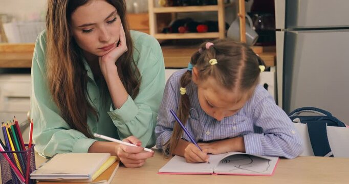 Bored mother looking at her daughter doing homework on the table.