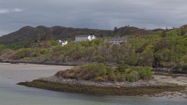 Drone tracking in a slow orbit above the Morar River, revealing the historic Morar Hotel perched on a green hillside in the Scottish Highlands.