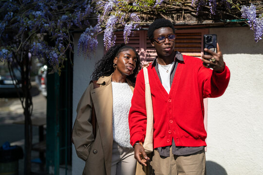 Stylish african american man and woman students pose for selfie outdoors on street together, positive relaxed black male and female friends take picture on smartphone against blooming Wisteria.