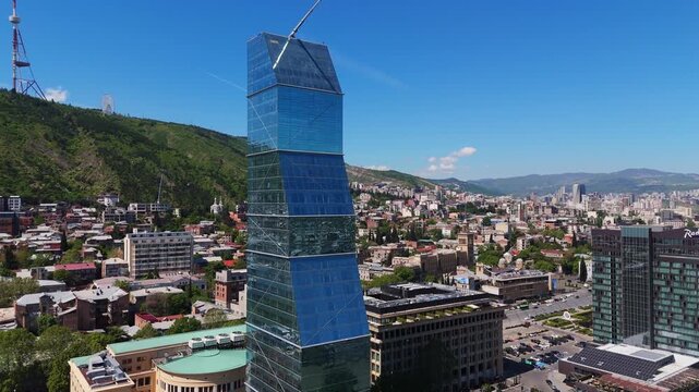 drone orbit around the Biltmore Hotel skyscraper in Tbilisi, Georgia, capturing a close-up of its glass facade and modern architectural design against the city skyline