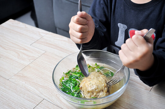 Close up shot of an Asian child holding a spoon and fork ready to eat a giant bowl of traditional Indonesian bakso meatball soup on a wooden table