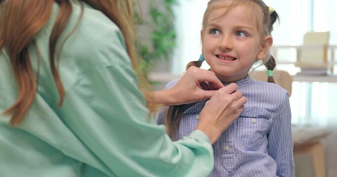 Mother buttoning her daughter shirt and helping her get ready for school.