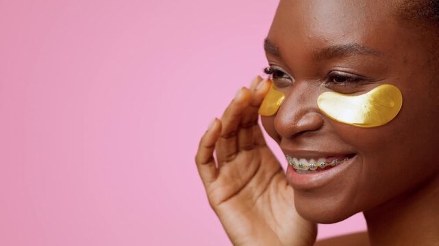 Smiling Young Black Woman Applying Golden Under Eye Patches On Her Face, Enjoying Skincare Routine At Home. Indoor Setting Focused On Beauty And Self-care, Concept Of Healthy Skin Care