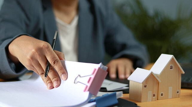 Real estate agent reviewing property documents and mortgage paperwork with model houses on office desk. Home buying process, housing investment, contract management, loan approval and property 