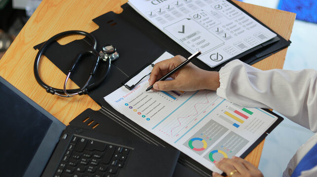 Healthcare professional analyzing medical reports and performance data on office desk with stethoscope and laptop. Healthcare analytics, patient records, clinical audit, medical statistics 