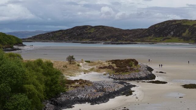 Telephoto drone flight tracking slowly backward over Morar Beach, capturing rocky outcrops, sand dunes, and the vast tidal estuary in the Scottish Highlands.
