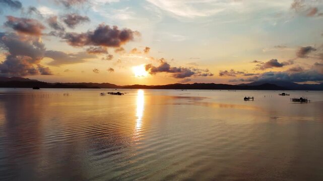 Aerial drone view of a glassy tidal bay at golden sunset with silhouetted bamboo fish traps and mountain ridges reflected in still shallow water, Taytay, Palawan, Philippines.
