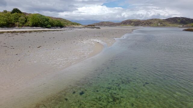 Low-altitude drone shot tracking sideways over the crystal-clear turquoise waters of the Morar River and the wide sands of Morar Beach.