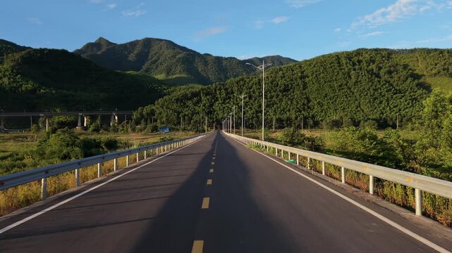 Low-altitude drone shot tracks steadily forward above an empty asphalt road equipped with guardrails and lampposts, heading toward a tunnel entrance at the base of lush green mountains under a clear b