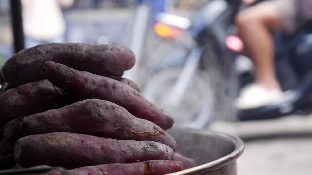 Sweet potatoes steam inside a metal container at a local outdoor market near a busy street. Traditional cooking.